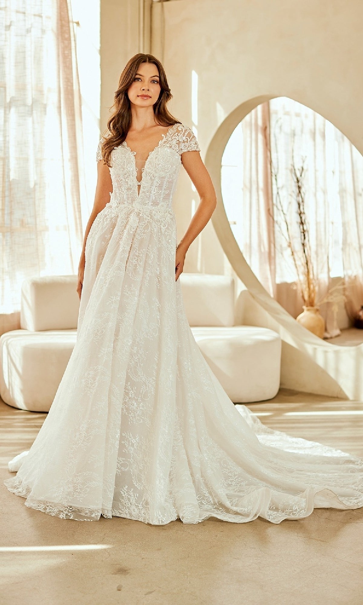 A woman stands indoors wearing the Amelia Couture 2142 long wedding dress with a lace A-line silhouette, corset bodice, cap sleeves, and a long train. Neutral furniture and sheer curtains are visible in the background.